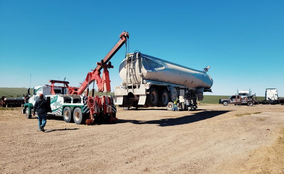 A large tow truck lifting a tanker truck on a dirt lot under a clear blue sky.