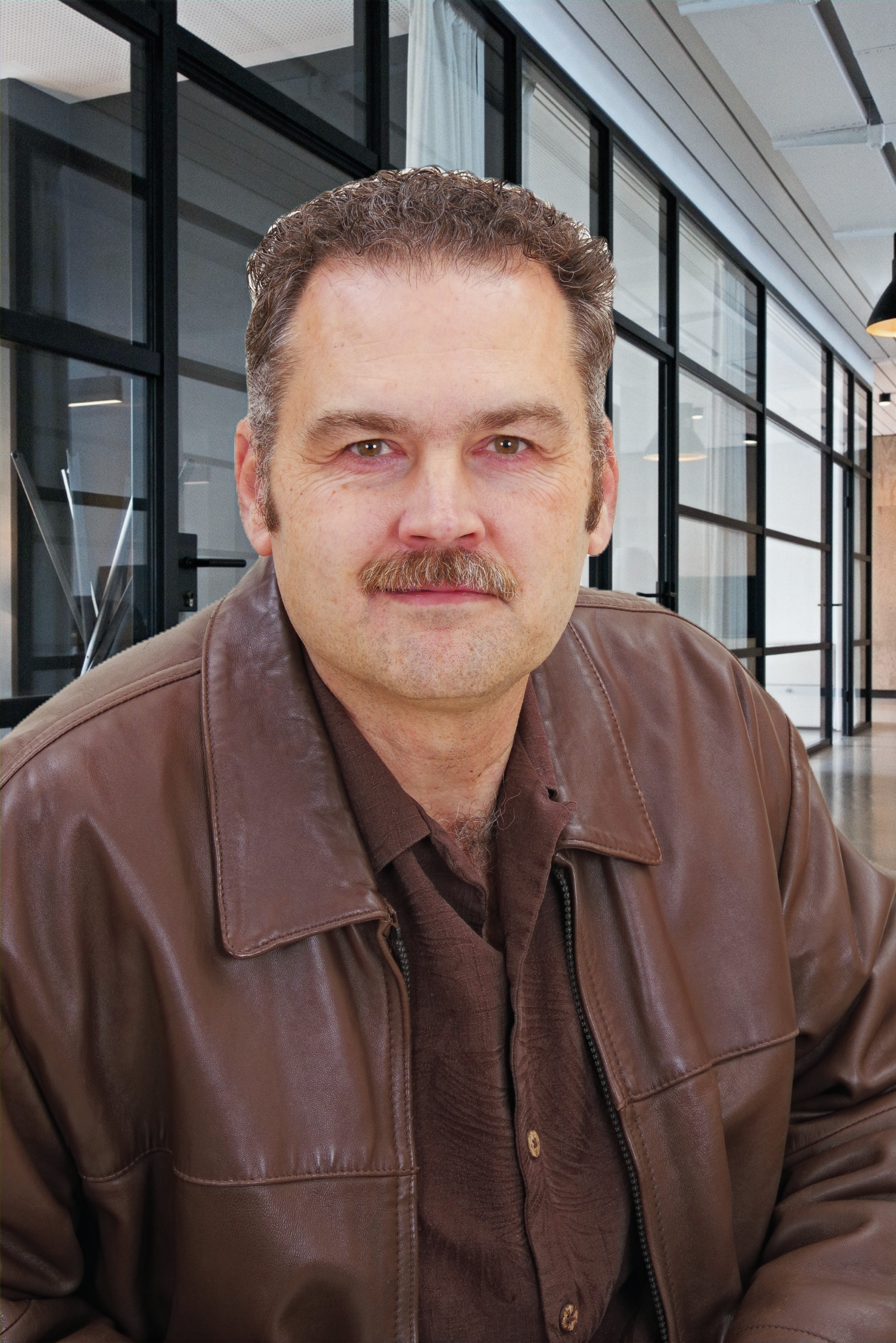 A man with a mustache is wearing a brown shirt and smiling.