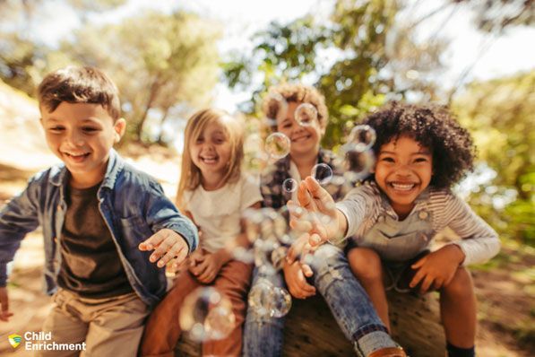 Four children sit outdoors, smiling and reaching toward floating bubbles in a sunlit, natural setting.