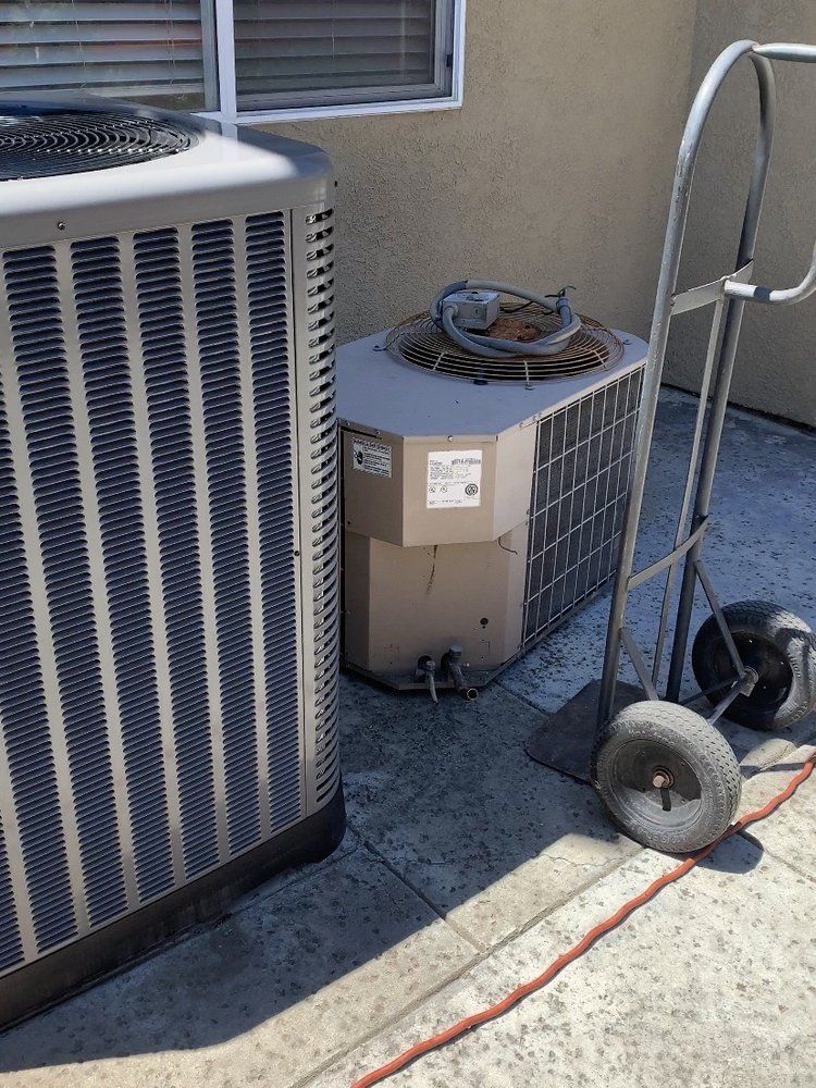 Two outdoor air conditioning units beside a house wall, with a hose and small cart on the ground.