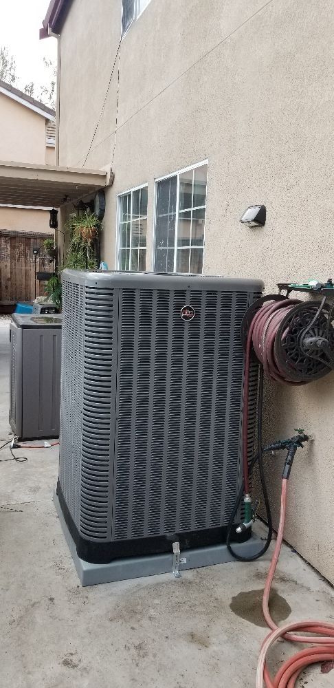Large gray outdoor HVAC unit beside a beige house wall, near a window and coiled hose.