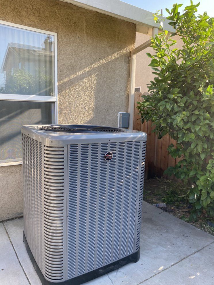 Outdoor air conditioning unit beside a house wall and window, next to a wooden fence and shrub.