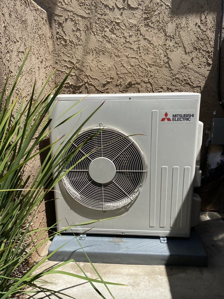 White Mitsubishi electric outdoor AC unit beside a beige stucco wall, partly hidden by plants.