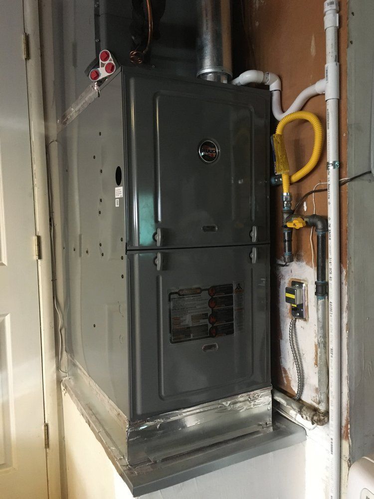 Dark gray furnace in a narrow utility closet with exposed pipes and a yellow hose on the wall