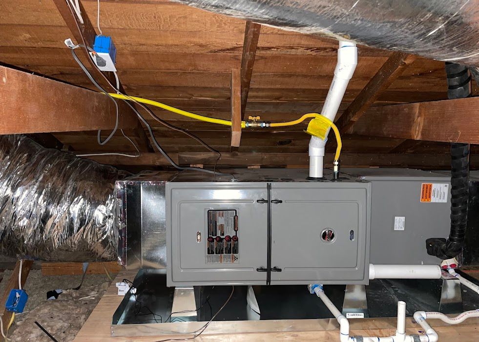 Gray HVAC unit in an attic with insulated ductwork, white vent pipe, and exposed wooden rafters.