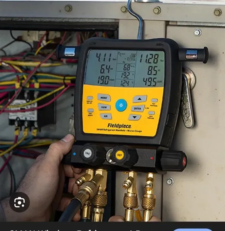 Yellow Fieldpiece HVAC gauge manifold with hoses connected, held in front of electrical equipment.