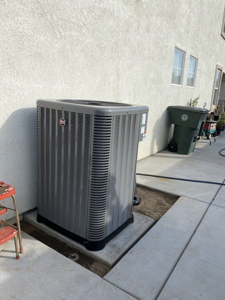 Outdoor air conditioning unit beside a building wall on a concrete patio, with trash bins nearby