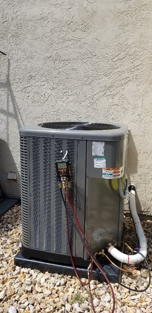 Outdoor gray air conditioner unit beside a beige wall and white hose on gravel.