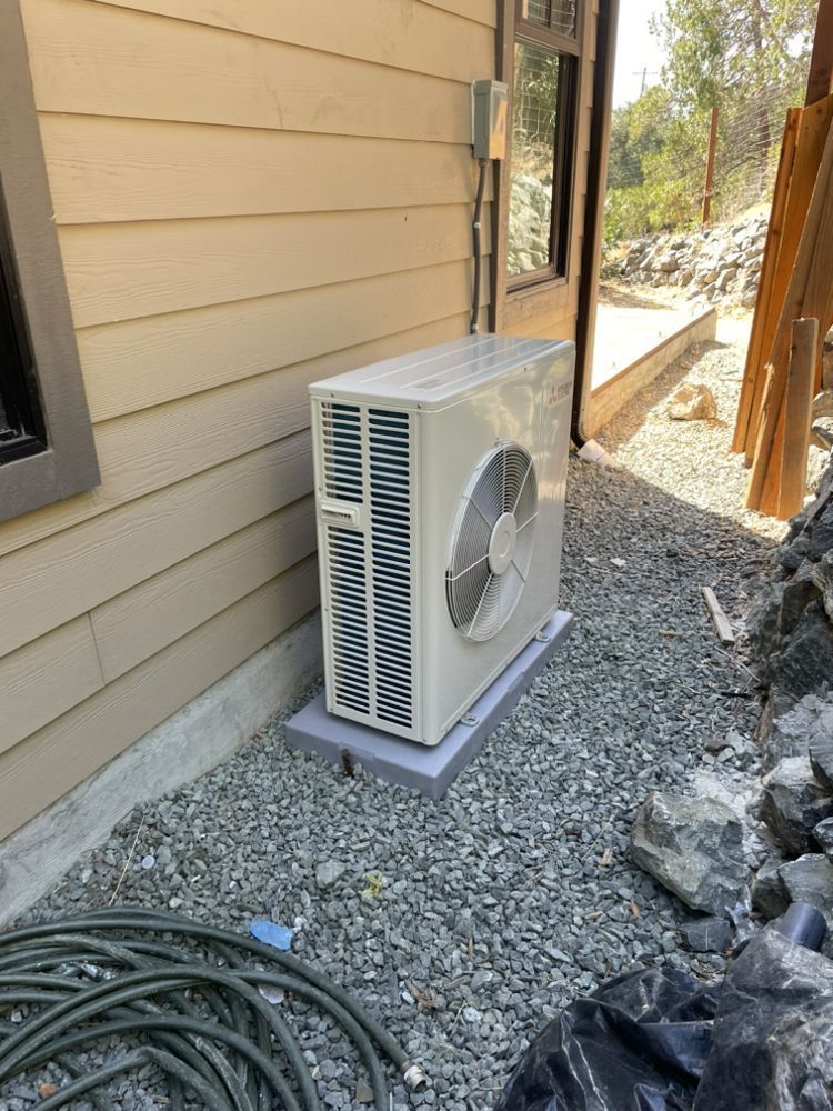 Outdoor mini-split AC unit beside a beige house wall on a gravel path, with garden hose nearby