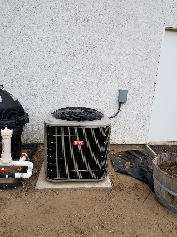 Outdoor air conditioning unit beside a white stucco wall, with dirt ground and nearby utility equipment