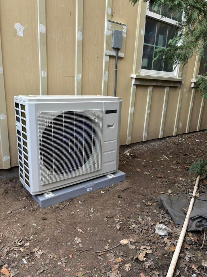 Outdoor air conditioner unit beside a tan house wall near a window, on bare dirt and leaves.