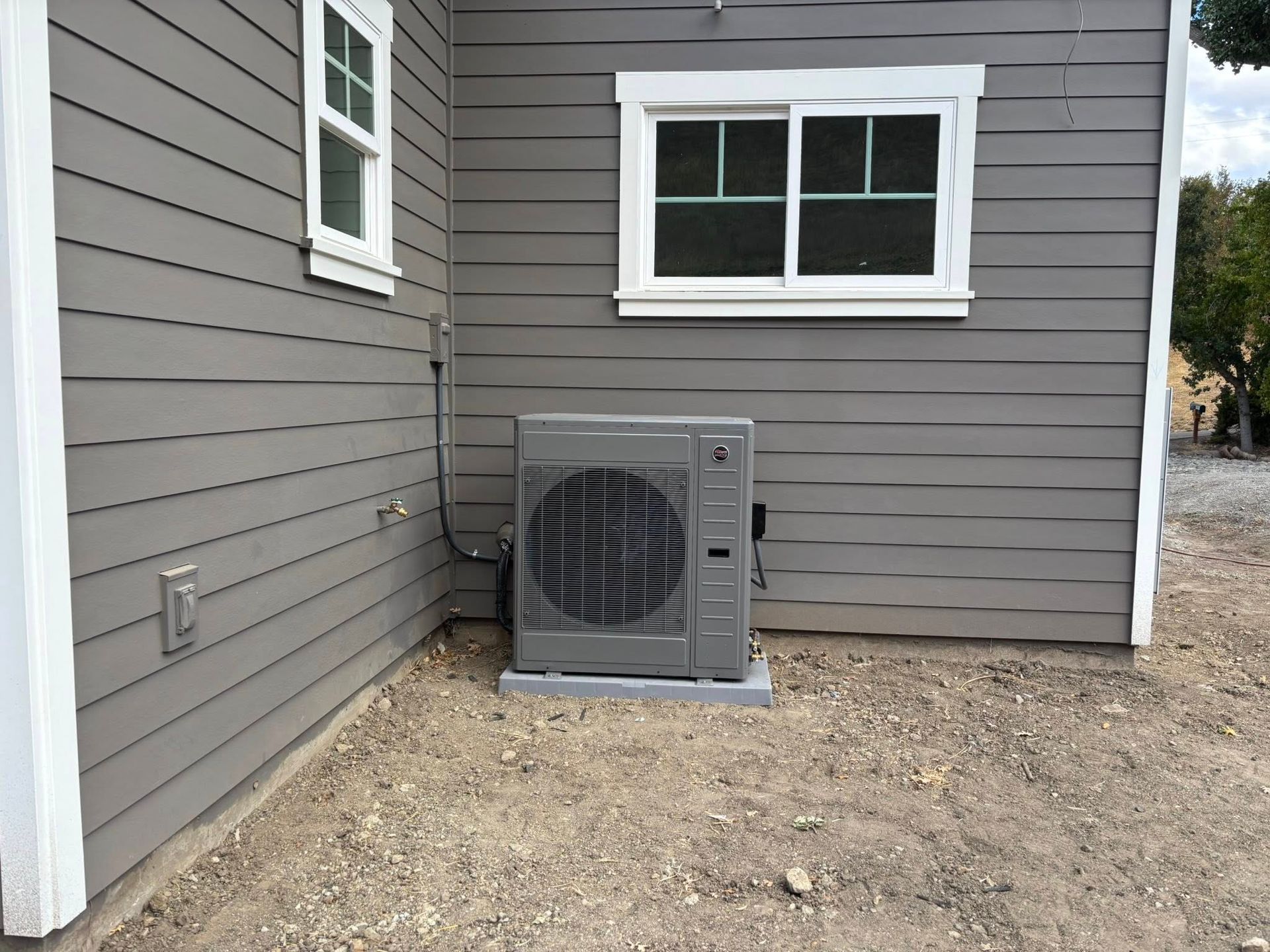 Exterior HVAC unit beside a gray house with white-trimmed windows on a gravel patch