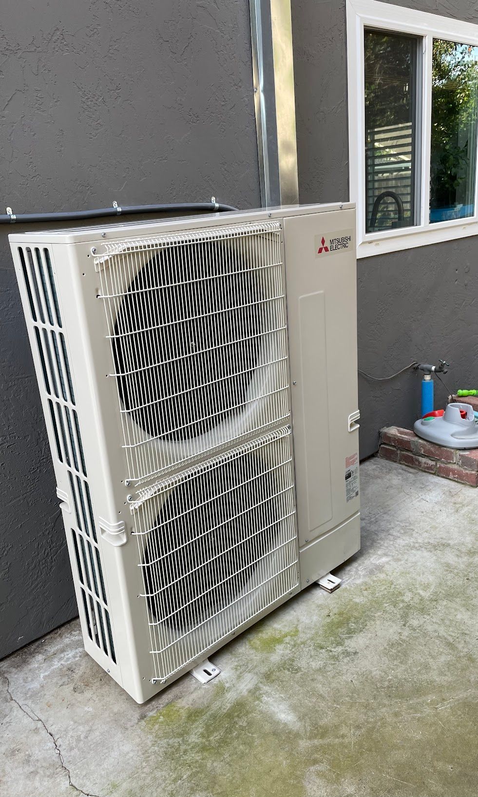 Two outdoor HVAC units beside a gray house wall and window, with a small patio item nearby.