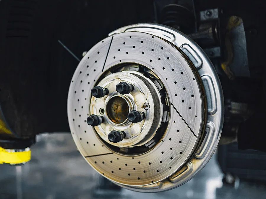 Close-up of a car's drilled and slotted brake rotor, mounted on the hub, with a black background.