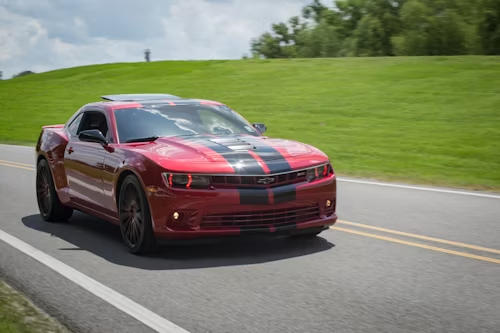 Red Chevy Camaro with black stripes driving on a road next to a grassy hill.