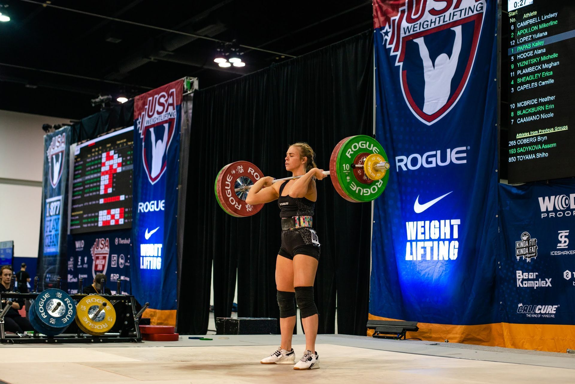 A weightlifter at a USA Weightlifting competition stands with a barbell across her chest, in front of a crowd and banners.