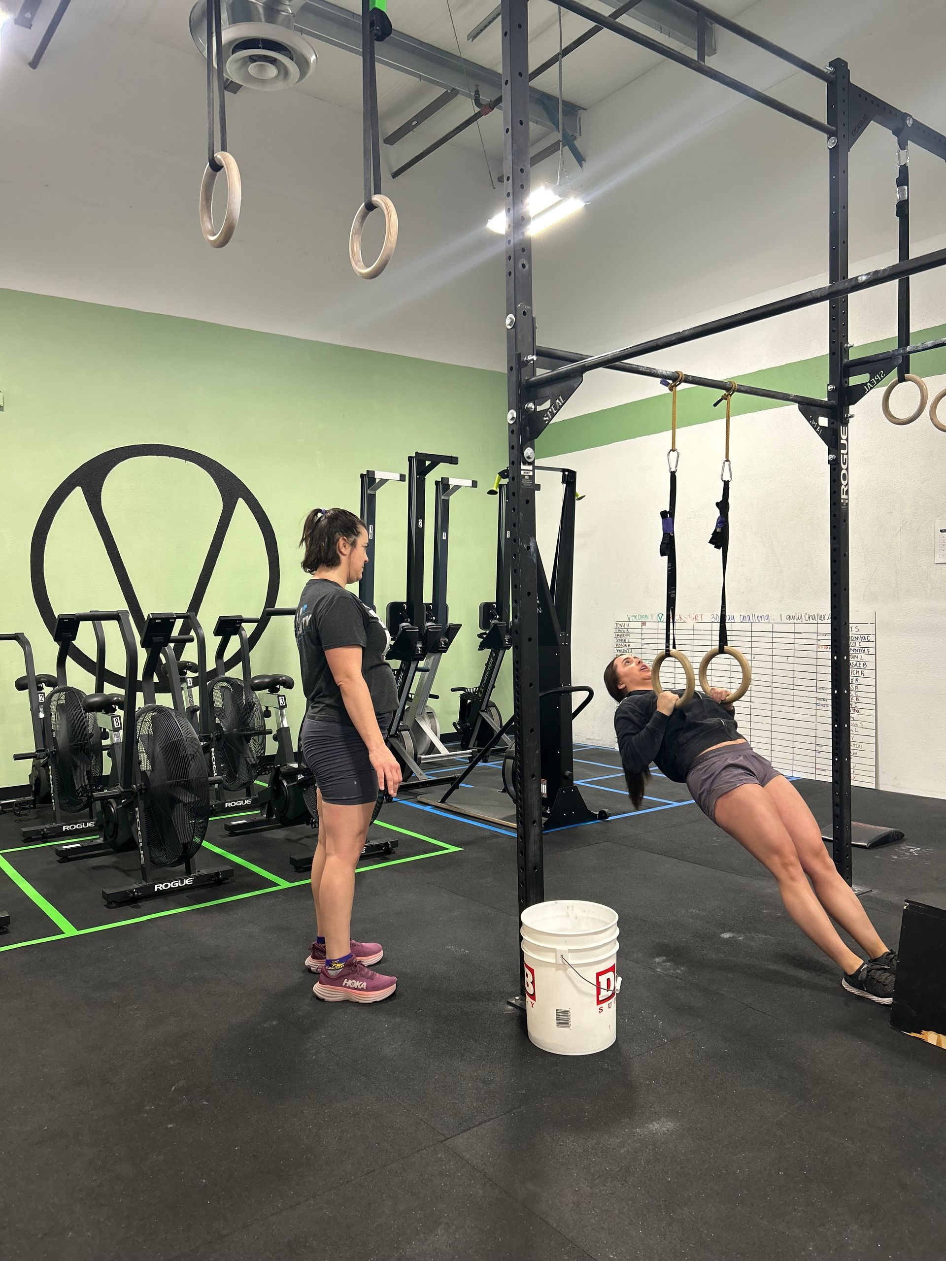 Woman doing a ring row exercise while another watches at Verdant CrossFit in Boise. Black rig, green wall, and gray floor.