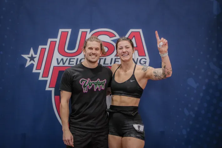 Man and woman smiling, standing in front of a USA Weightlifting banner. Woman raises a finger.