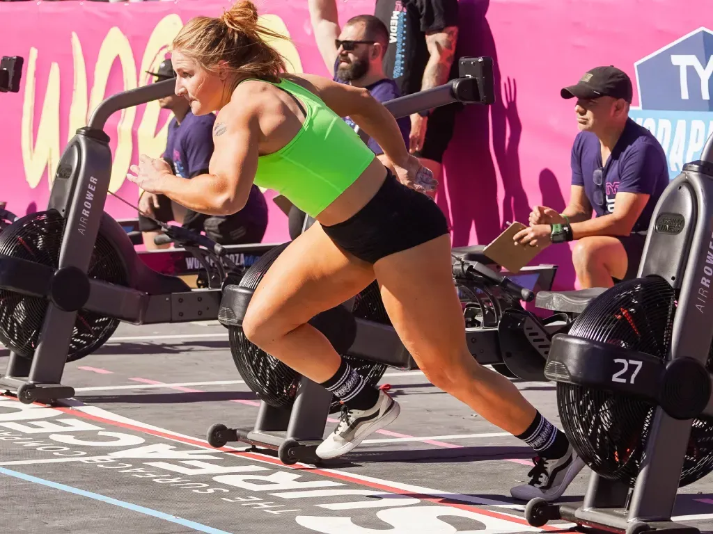 Athlete running toward air bike in a competition. Wearing a neon green top, black shorts, setting is outdoors.