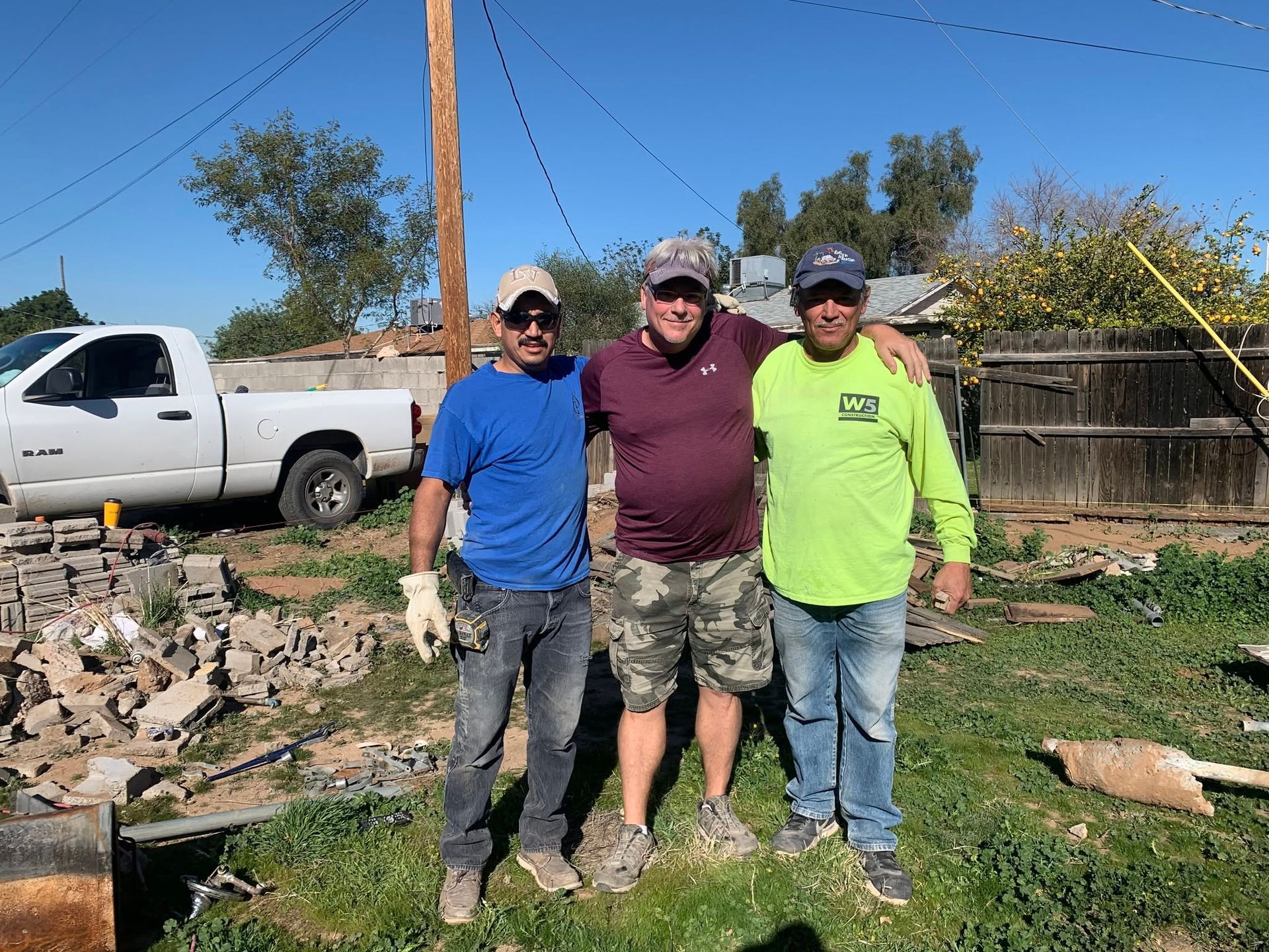 Three people standing outdoors in a yard beside a white truck and rocks, smiling at the camera.