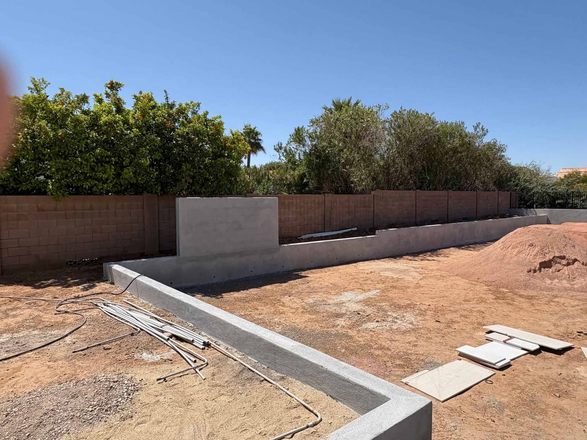 Concrete foundation and retaining walls under construction in a dry backyard with a brick fence and trees.