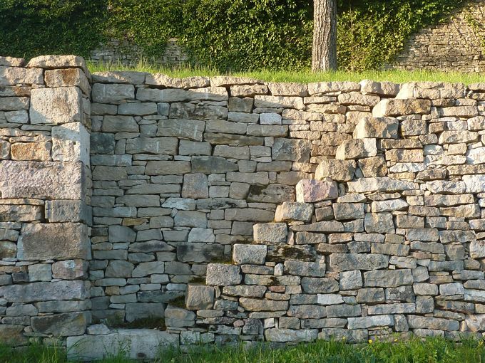 Stone retaining wall with a recessed section, sunlit grass and trees above it.