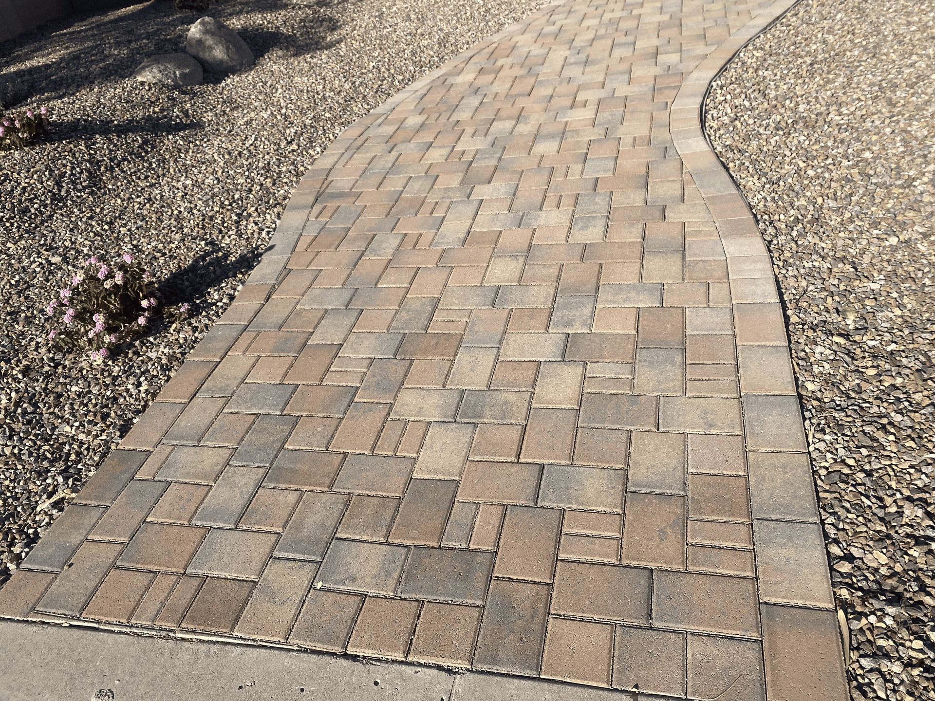 Curved tan brick walkway bordered by gravel and rocks in a landscaped area