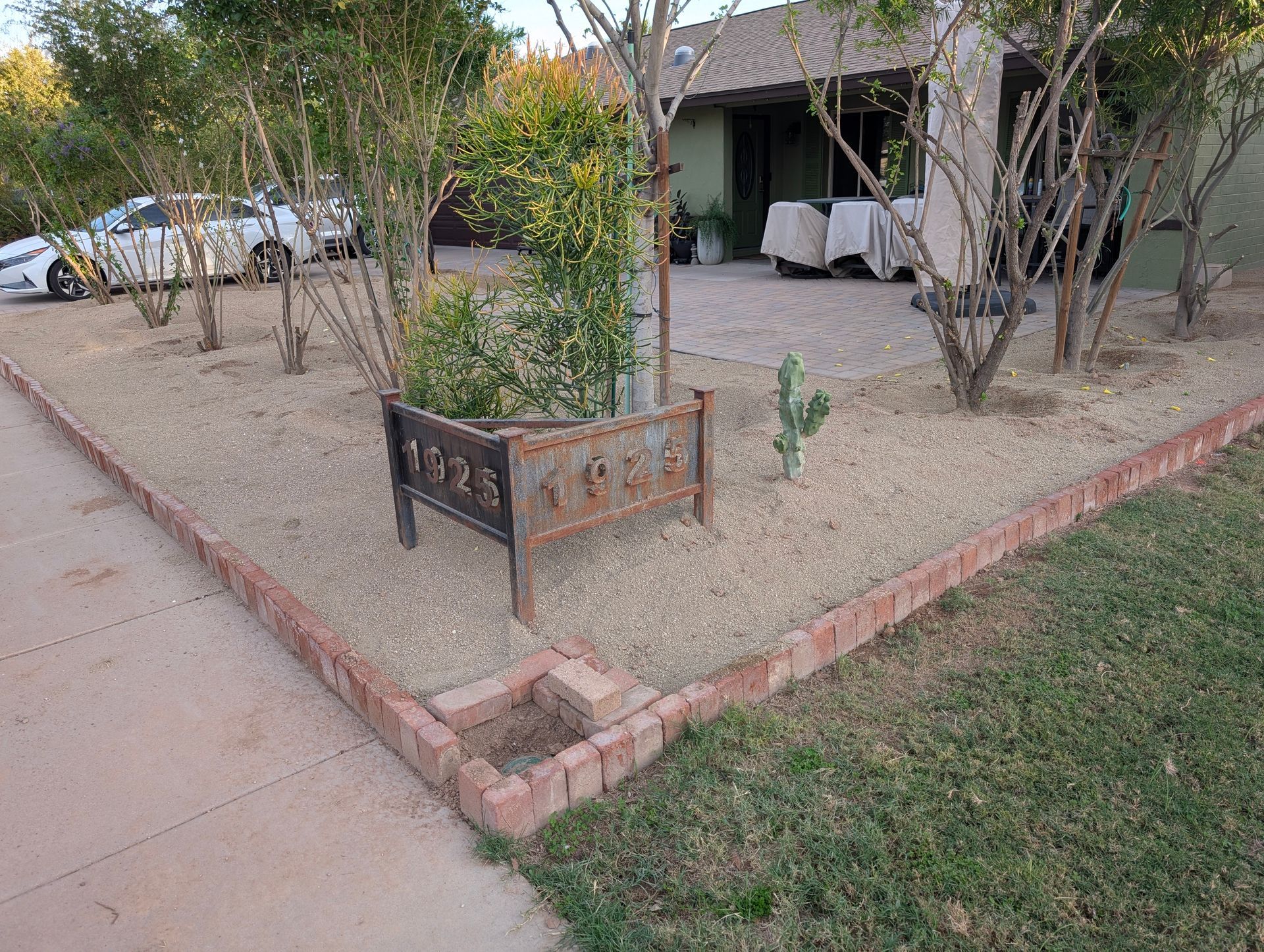 Front yard with a small fenced planter, trees, and a covered patio beside a driveway.