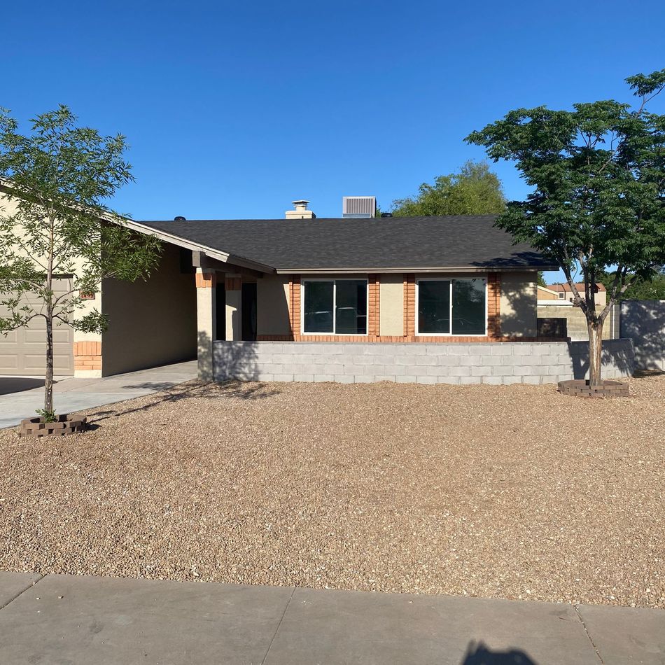 Single-story house with stone facade, dark roof, gravel front yard, and trees under a clear blue sky
