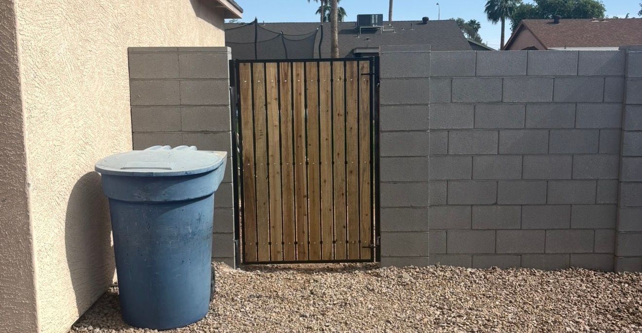 Backyard side gate with gray block wall, wooden gate, and blue trash bin on gravel.