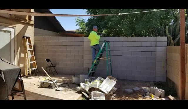 Worker in neon jacket on a ladder building a cinder block wall in a backyard, with debris on the ground