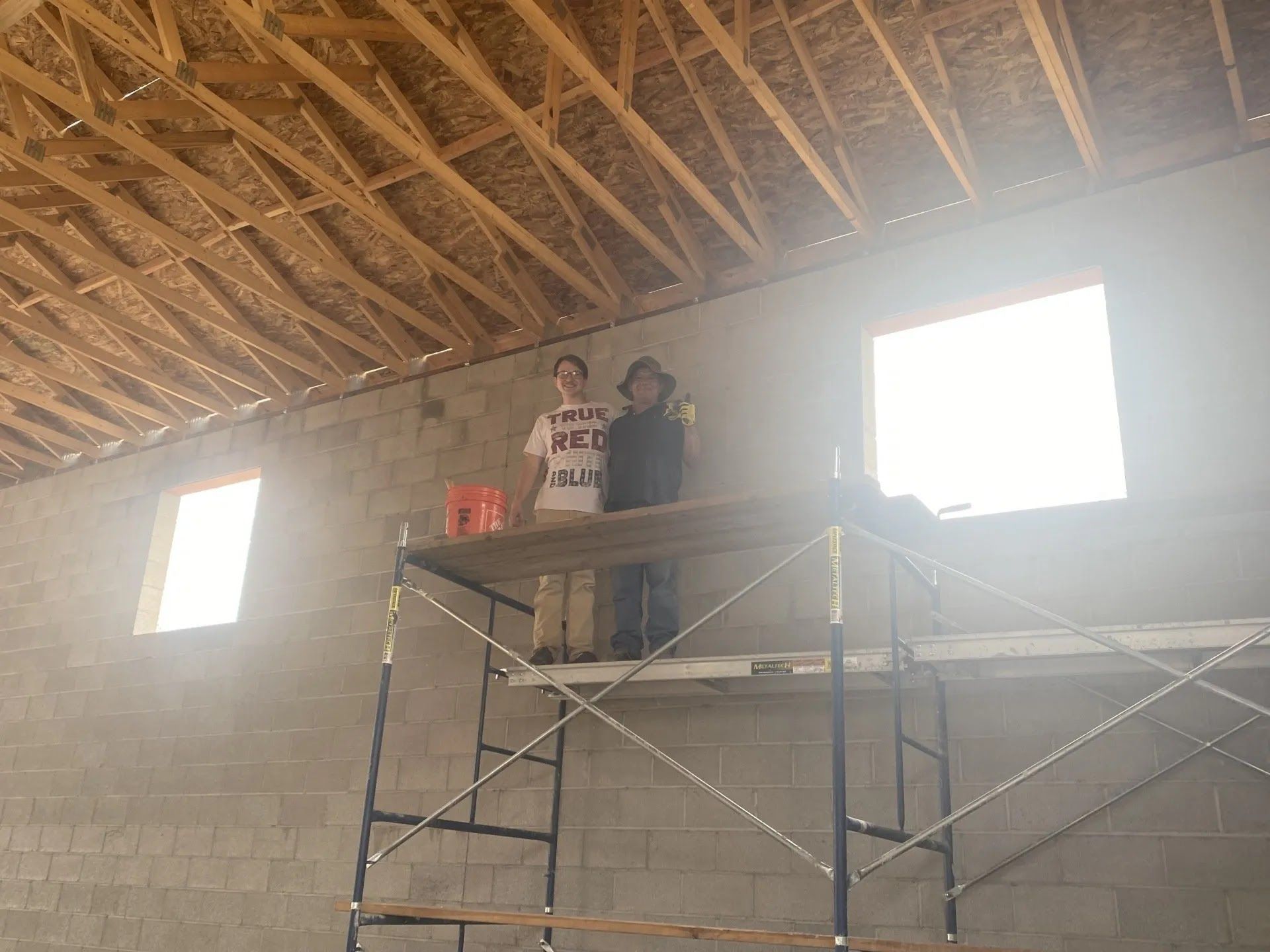 Two people standing on scaffolding inside a cinderblock building under construction, with exposed roof trusses.