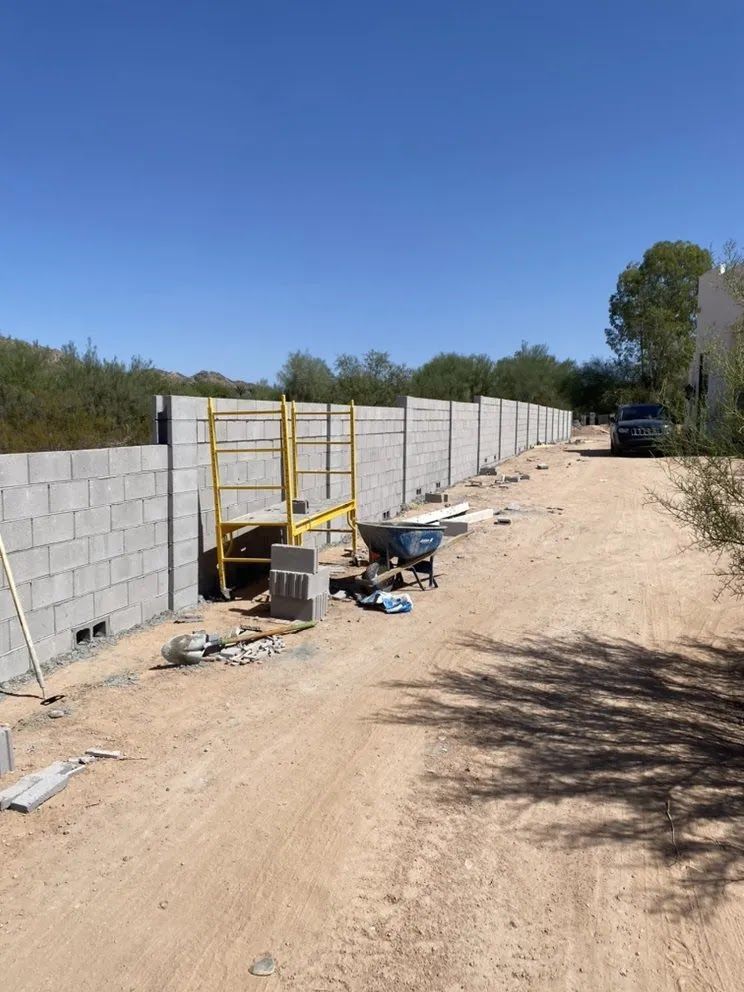 Construction site with a long cinder block wall, yellow scaffolding, and tools along a dirt road under a clear blue sky