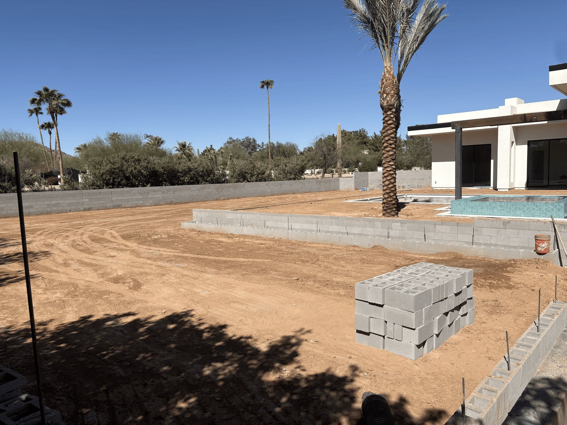 Desert yard with dirt ground, stacked cinder blocks, palm tree, and a low house under construction