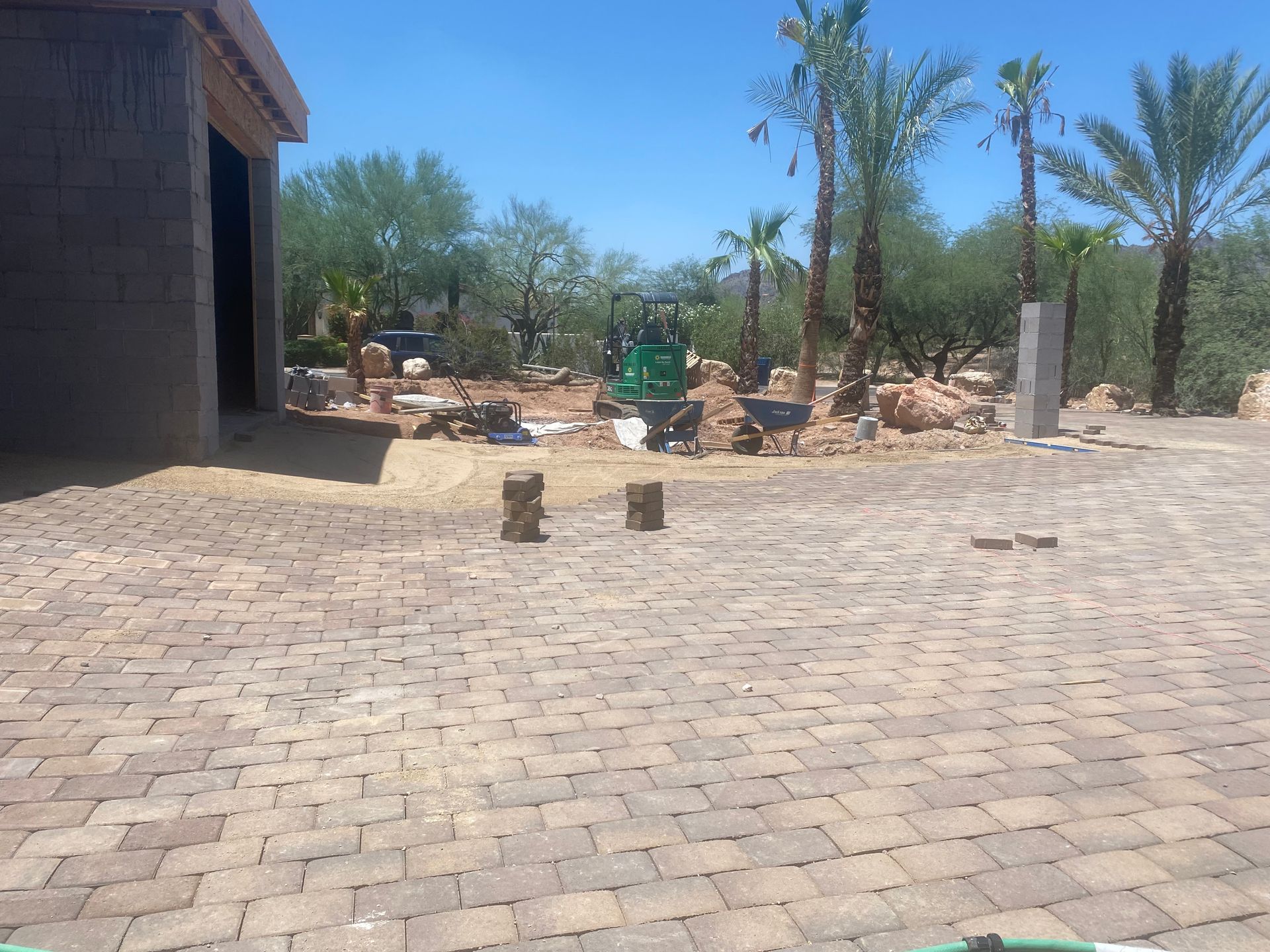 Paved courtyard with a small green tractor, palm trees, and stacked materials under a clear blue sky