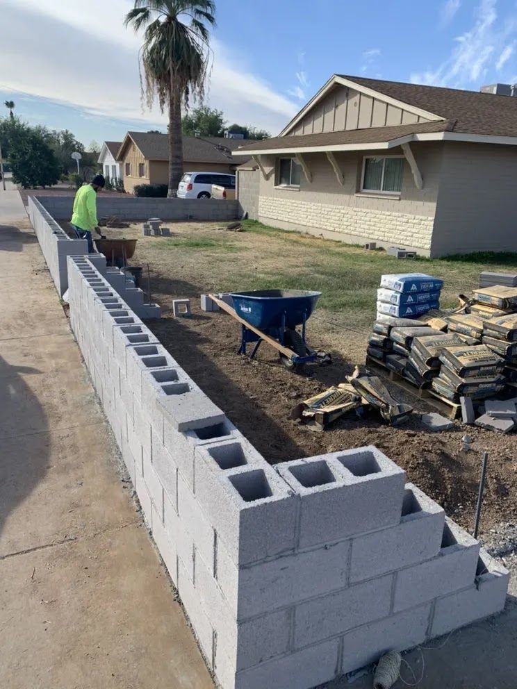 Workers building a cinder block wall along a sidewalk in a residential yard with tools and materials nearby