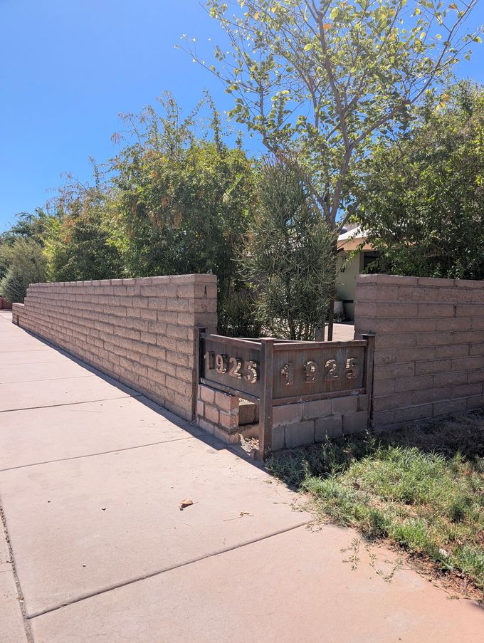 Sidewalk beside a tan block wall with a small wooden gate and trees behind it under a blue sky