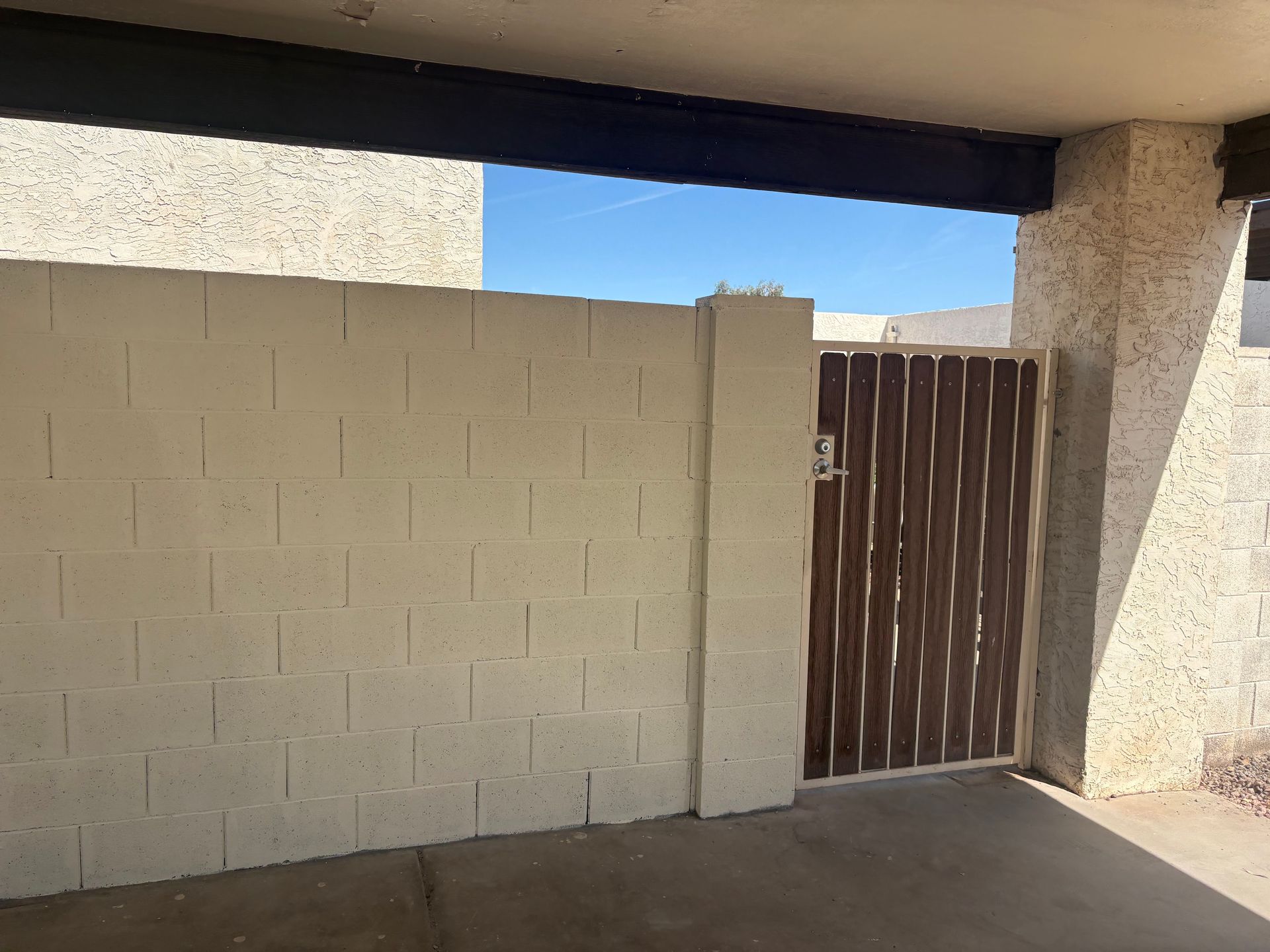 Beige stucco wall and wooden gate under a shaded patio, with blue sky above