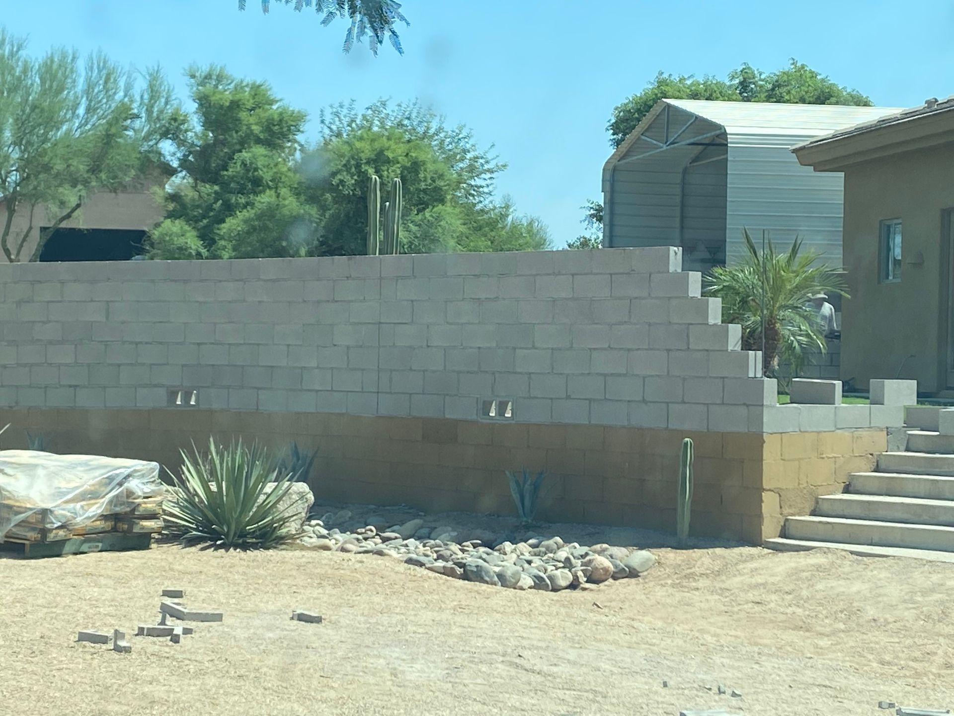 Block wall in a desert yard beside stairs and a house, with rocks, plants, and blue sky.