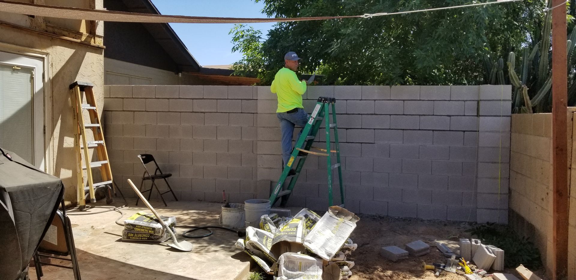 Worker in neon shirt on ladder building a cinder block wall in a backyard.