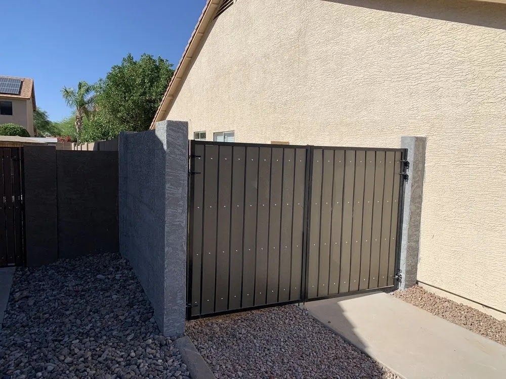 Closed dark gray metal gate beside a beige stucco house in a gravel driveway.