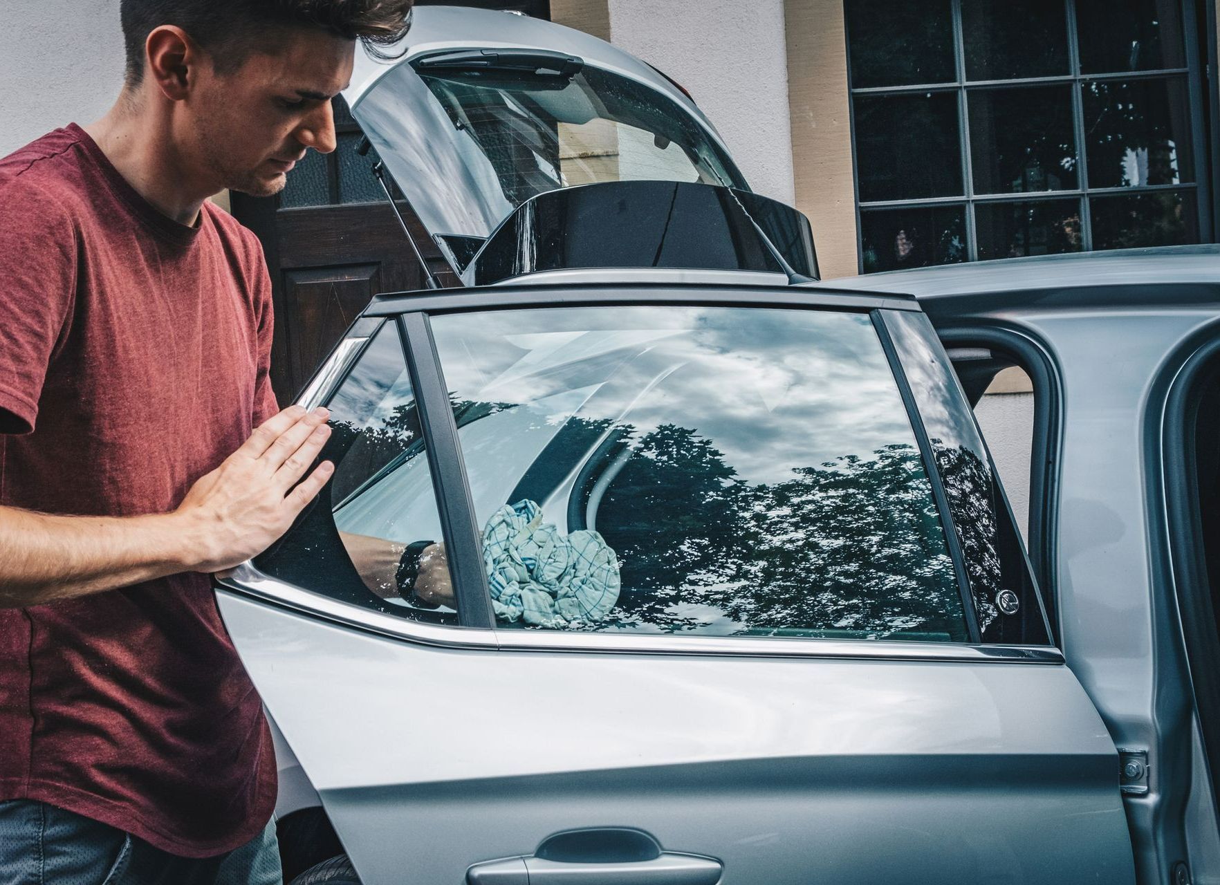 A man in a red shirt cleans the window of a silver car with a cloth, outdoors.