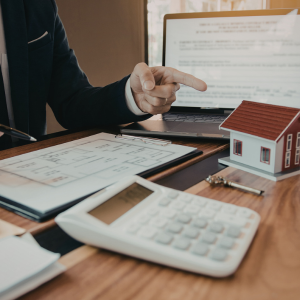 Person points at documents beside a calculator, laptop, and small house model on a desk.