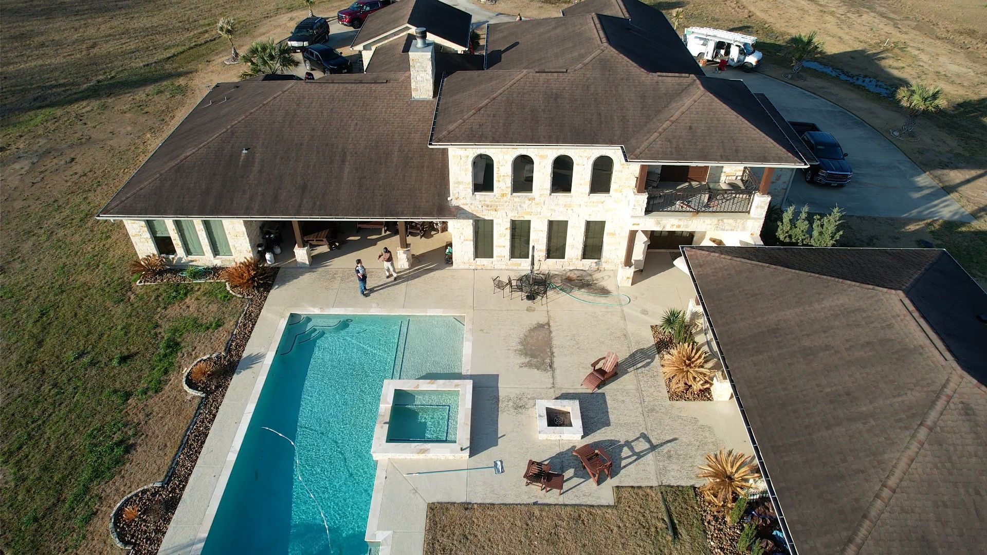 Aerial view of a large house with a pool and patio. Cars parked in the driveway, brown roof.