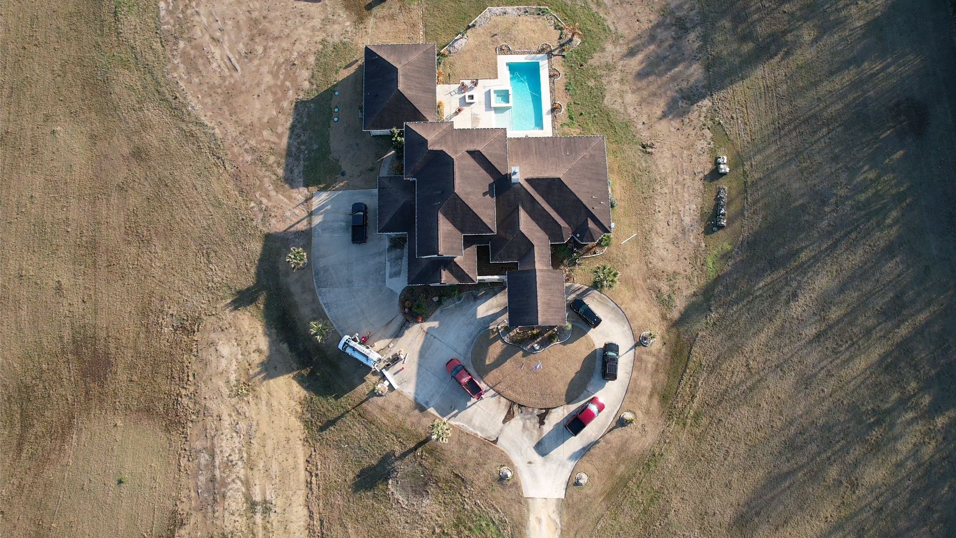 Aerial view of a house with a pool, surrounded by dry, grassy land. Cars are parked in front.