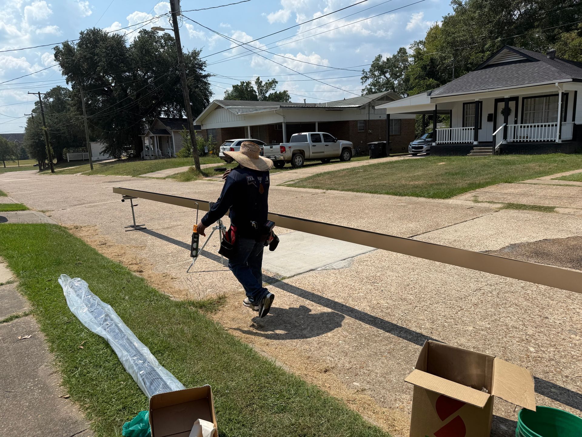 Person carrying a long, brown beam on a gravelly sidewalk near houses and a truck on a sunny day.