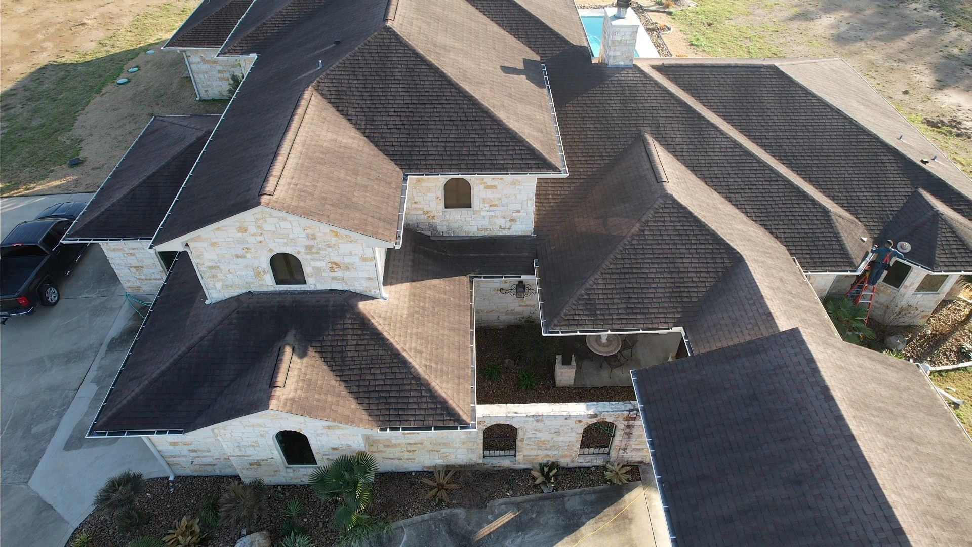 Overhead view of a stone-walled house with a dark shingle roof, surrounded by trees and a driveway.