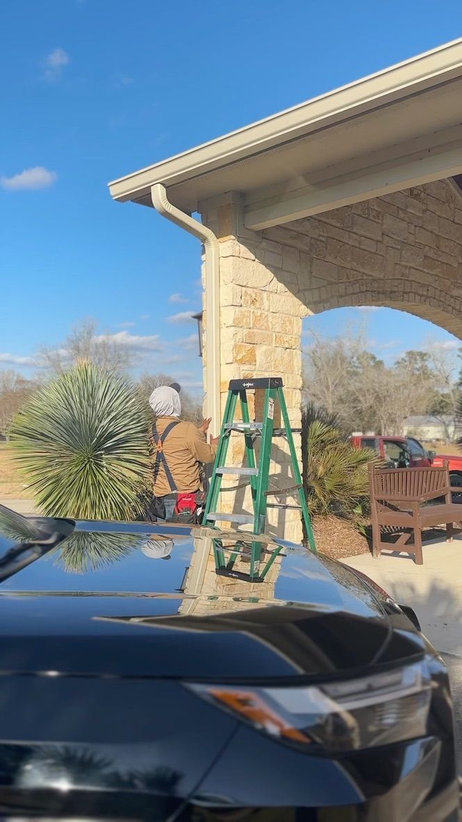 Person on ladder near building cleaning gutters, sunny day.