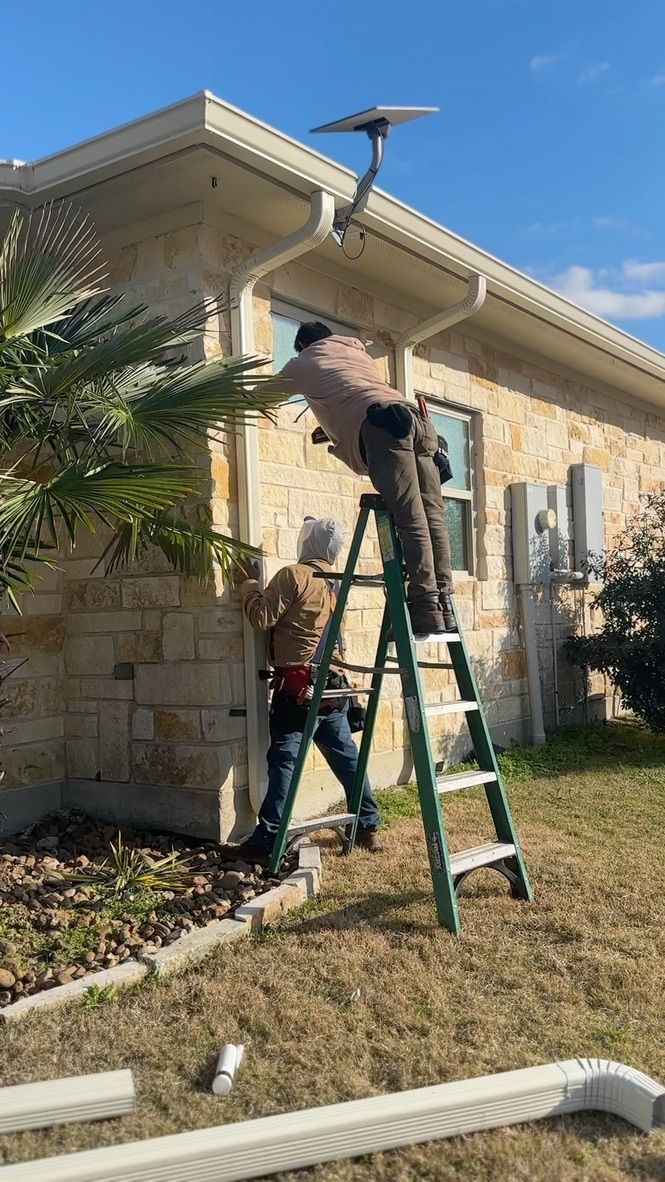 Two workers install satellite dish near the roof of a house; one on ladder, one on ground.