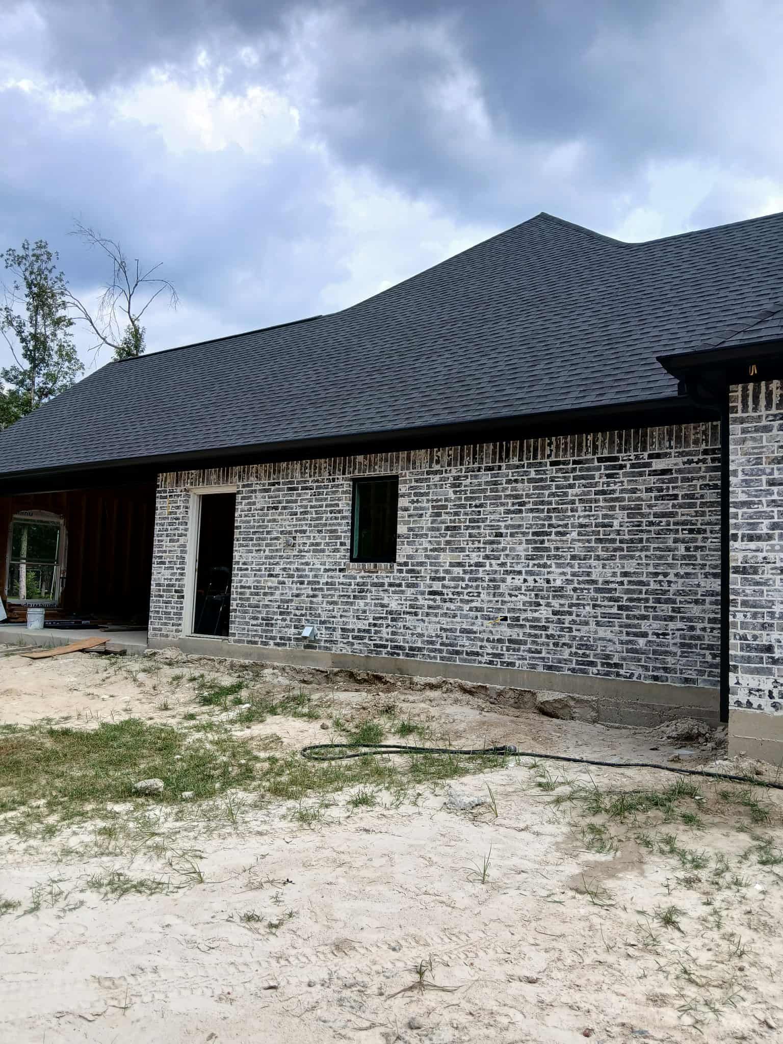 Brick house under construction with a dark shingle roof and a cloudy sky.